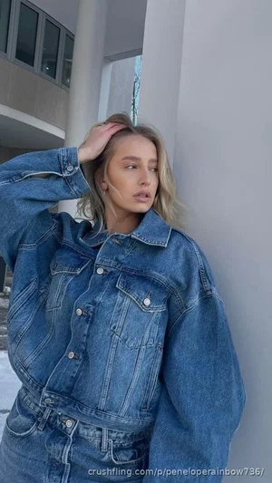 Woman in denim jacket and jeans leaning against a wall outdoors.