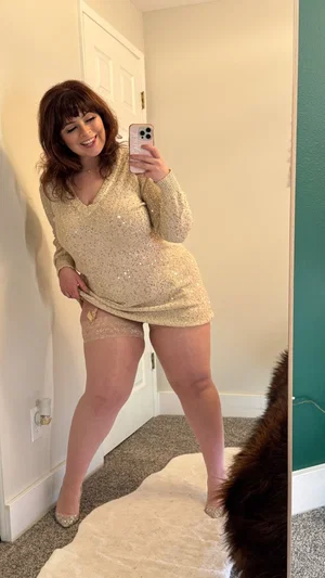 Woman in sparkly long-sleeve top and lace shorts takes mirror selfie in bedroom.