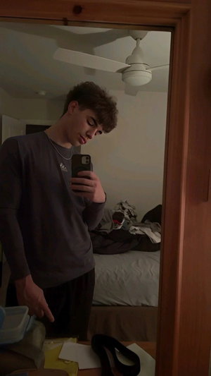 Young man in gray long-sleeve shirt and dark pants takes mirror selfie in bedroom.