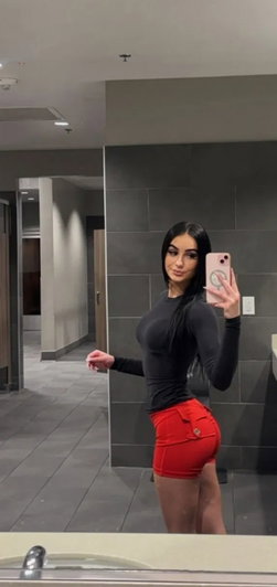 Dark-haired woman in black top and red shorts taking bathroom mirror selfie.