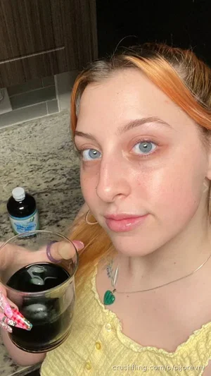 Woman with orange hair and blue eyes in yellow top holding drink in kitchen.