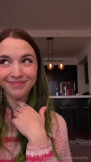 Woman with green-tipped hair in a pink sweater, smiling indoors.