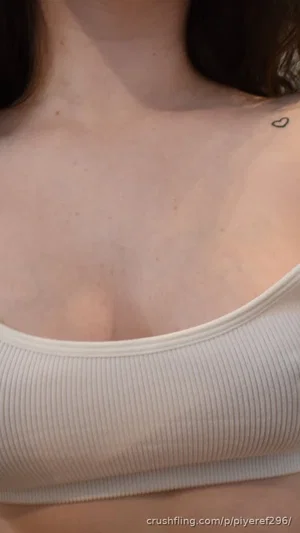 Woman with dark hair wearing white ribbed tank top, small heart tattoo on collarbone.