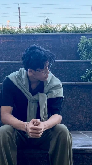 Man with dark hair and glasses sitting outdoors, wearing black shirt and olive pants with scarf.