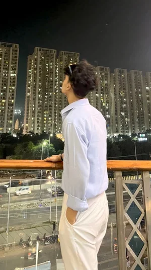 Man in light blue shirt and white pants standing on balcony at night, city buildings in background.
