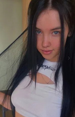 Woman with long black hair in white crop top, indoors, facing camera.