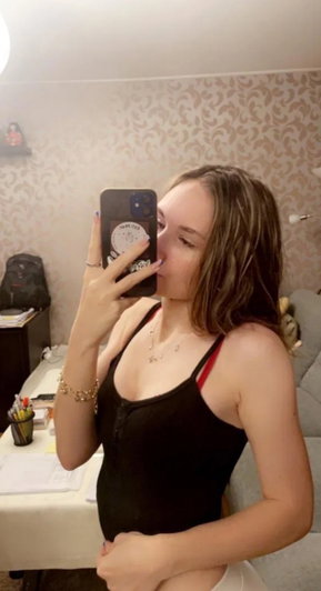 Brunette woman in black bodysuit taking mirror selfie indoors.