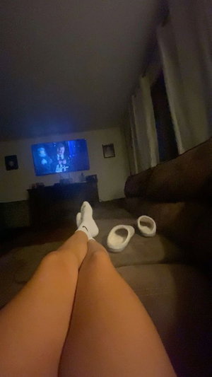 Person sitting on couch with legs extended, wearing white socks, TV on in background.