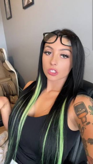 Woman with black and green hair, tattoos, wearing black top, indoors.