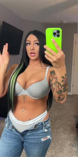 Woman with long black hair and green streaks in gray bra and jeans, taking mirror selfie indoors.
