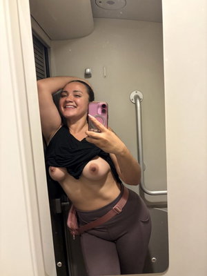 Curvy woman taking mirror selfie in bathroom with top lifted.