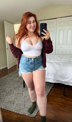 Red-haired woman taking selfie in bedroom wearing white top and shorts.