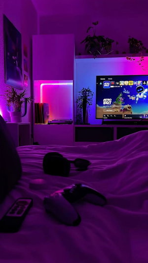 Bedroom with unmade bed, TV, and gaming controllers under purple lights.