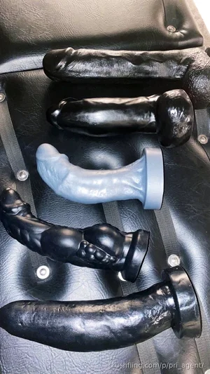 Five black and one light blue dildo toys arranged on a black leather surface.