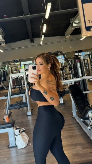 Woman in black workout outfit with tattoos takes mirror selfie in gym.