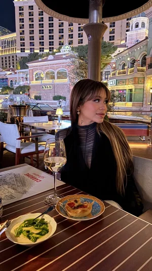 Woman with long hair in black sheer top sits at outdoor restaurant table with food and wine.