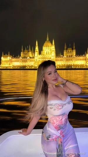 Woman in floral dress posing on boat at night with illuminated building in background.