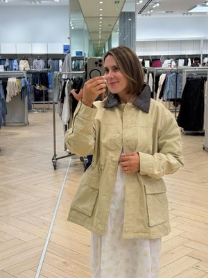 Woman with brown hair in beige jacket taking mirror selfie in store.