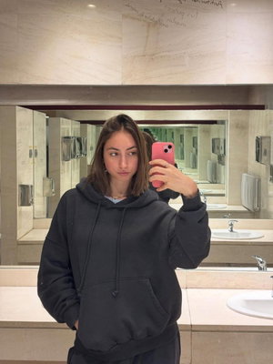 Woman in black hoodie taking mirror selfie in bathroom.