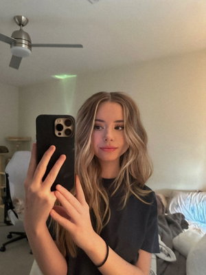 Woman with wavy blonde hair in black t-shirt takes mirror selfie in bedroom.