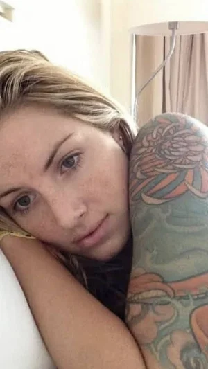 Woman with blonde hair and freckles resting head on arm, colorful tattoo on arm, indoor setting.