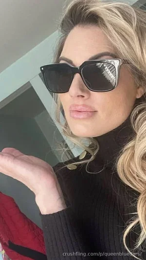 Blonde woman in black turtleneck and sunglasses, posing indoors.