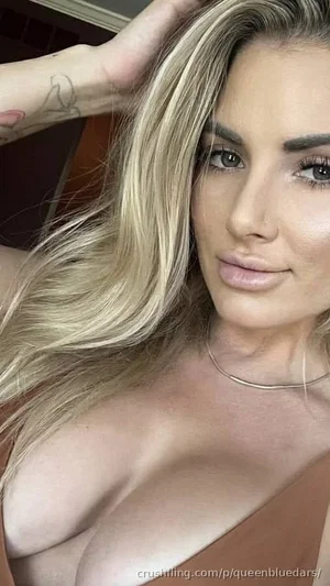 Blonde woman in brown top, posing indoors with hand in hair.