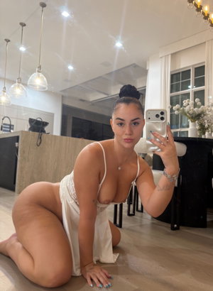 Woman in white lingerie posing on floor in modern kitchen, holding phone for mirror selfie.