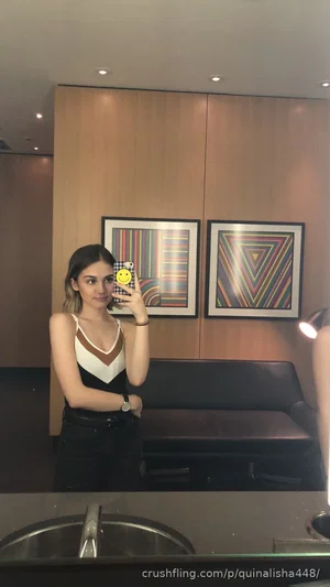 Woman in black and white top, dark pants, taking mirror selfie in modern room with art on wall.