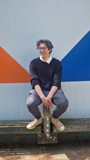Young man with glasses in sweater and pants sitting outdoors.