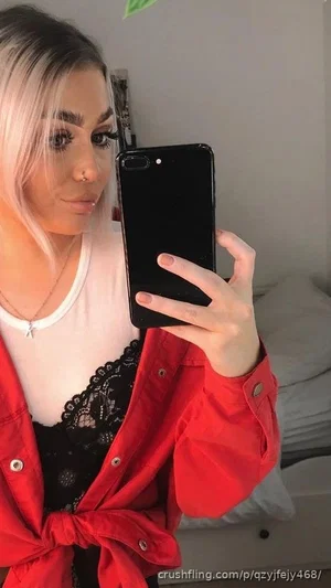 Woman with blonde hair in red jacket and black lace top taking mirror selfie in bedroom.