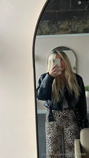 Blonde woman in black leather jacket and leopard print pants taking mirror selfie indoors.