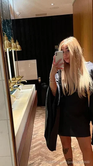 Woman with long blonde hair in black dress taking mirror selfie in bathroom.