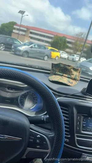 Car interior with steering wheel and dashboard, camouflage cap on dashboard, parking lot outside.