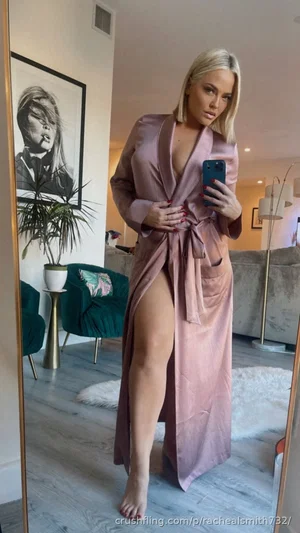 Blonde woman in a pink robe with a high slit, taking a mirror selfie in a modern living room.
