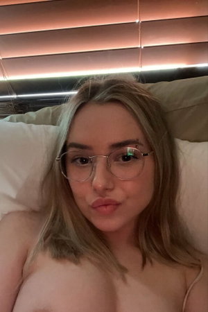 Woman with blonde hair and glasses lying in bed, bare shoulders visible.