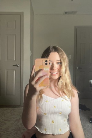 Woman with blonde hair in white crop top taking mirror selfie in bedroom.
