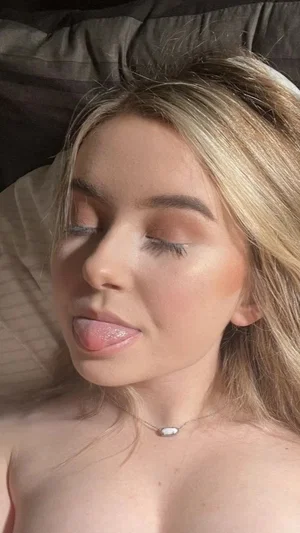 Blonde woman lying on bed, topless, wearing necklace, eyes closed, tongue out.