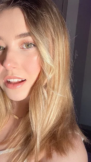 Blonde woman with green eyes, wearing a white top, indoors, close-up selfie.