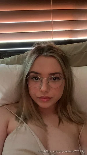 Woman with blonde hair and glasses lying in bed, wearing a light-colored top.