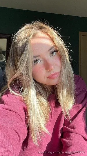 Blonde woman in a pink sweatshirt taking a selfie indoors.