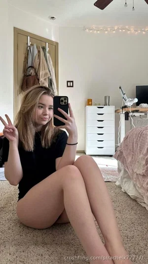 Woman with blonde hair in black top sitting on carpet in bedroom, holding phone for mirror selfie.
