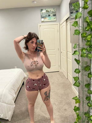 Woman in crop top and shorts taking mirror selfie in bedroom.