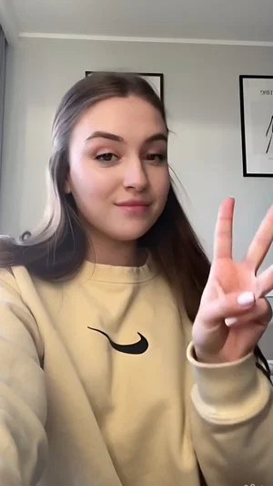 Woman with long brown hair in beige sweatshirt making peace sign indoors.