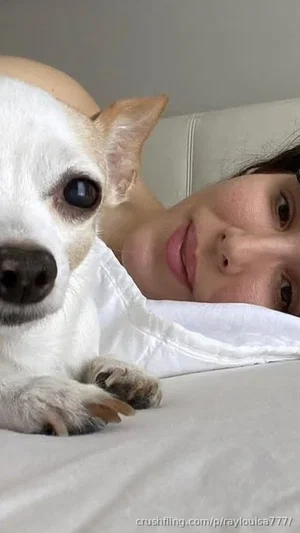 Woman with dark hair lying in bed with a small white dog, wearing a white top.