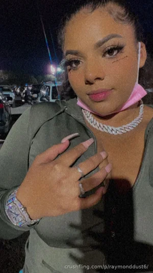 Woman in olive green top with pink face mask around neck, posing outdoors at night.