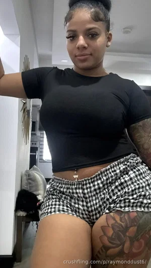 Woman in black crop top and checkered shorts, standing indoors with tattoos visible.