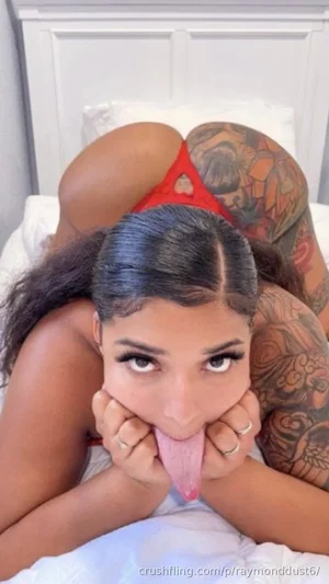 Woman with tattoos in red lingerie on bed, face close to camera, hands near mouth.