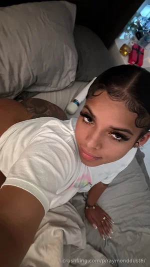 Woman with dark hair and makeup lying on bed in white t-shirt, tattoo visible on hip.
