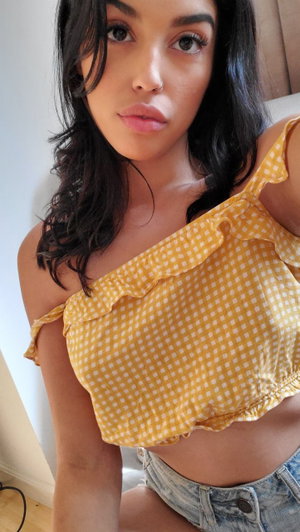 Woman with dark hair in yellow checkered top and denim shorts, sitting indoors.